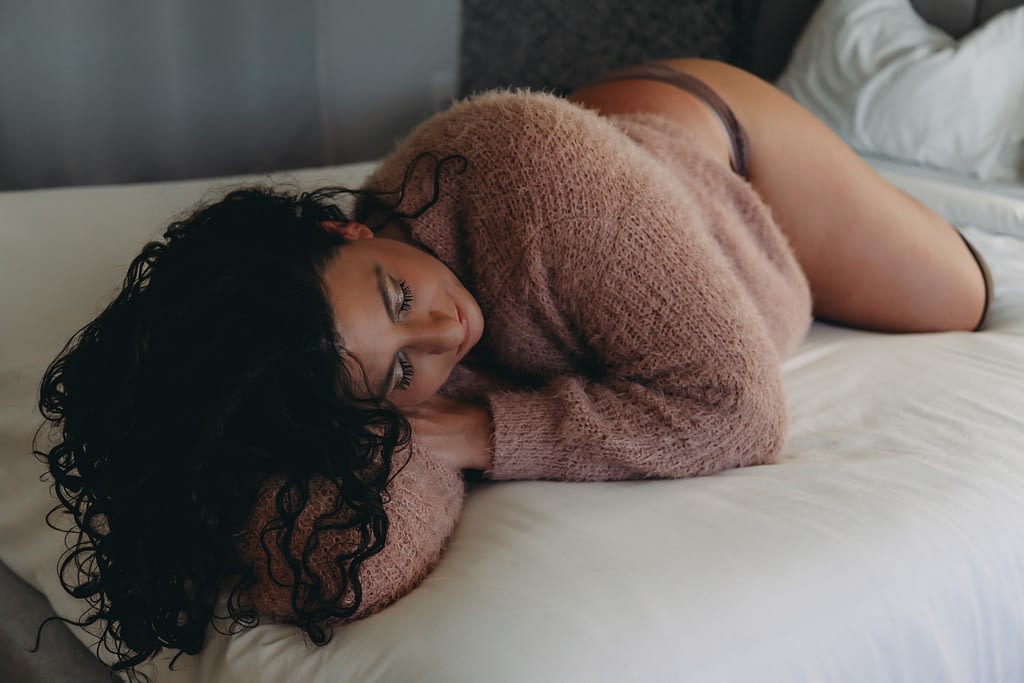 Middle aged woman laying on the bed peacefully, wearing only lace cheeky panties and a fuzzy pink sweater. Her curly brown hair spills over her arm. Photo by Embodied Art Boudoir. Body positive, body positive photography, fat positive, fat positive art, fat positive photography, colorado boudoir, denver boudoir, boulder boudoir, body positive colorado, haes, plus size photography, plus size art, women, beautiful, body positive art, big body art, you are beautiful, inclusive photography, inclusive art, celebrate all bodies, diverse photography