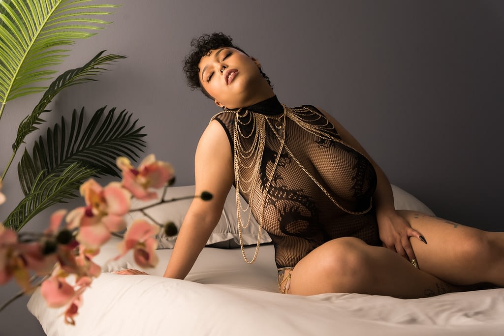 A woman wearing a black bodysuit and gold body chains lounging on a bed and caressing her leg. Photo by Embodied Art Boudoir. Denver boudoir photographer, boudoir photography, boudoir, colorado boudoir, photoshoot prep, boudoir inspiration, boudoir inspo, boudoir ideas, affirmation, positive, body positive, body neutral, confidence, confidence boost, mirror work, journaling, self care, self love, affirmations, self acceptance