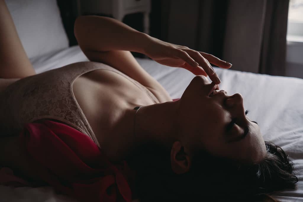 Woman lying on bed with eyes closed, touching lip wearing a pink bodysuit. Photo by Embodied Art Boudoir. Dark and moody, black and white photography, intense images, high intensity, memorable, sensual photography, sensual boudoir, colorado boudoir, denver boudoir, boulder boudoir, colorado springs boudoir, boudoir ideas, boudoir poses, boudoir inspiration, photography inspiration, romantic boudoir, bridal boudoir