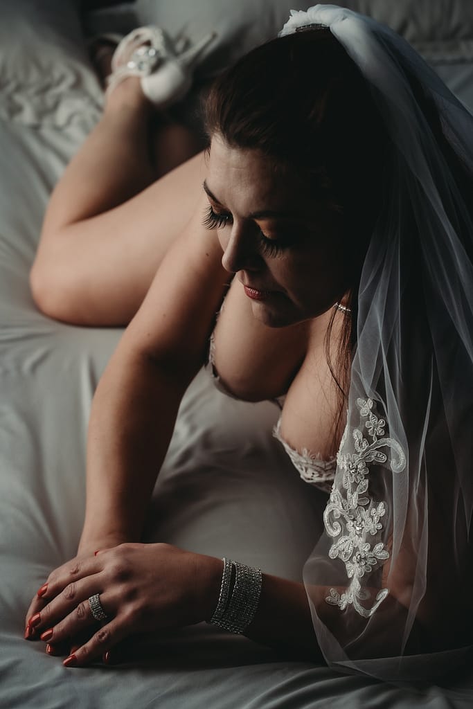 Bride dressed only in white lingerie, white heels, and a white veil with lace details lounging on the bed. Photo by Embodied Art Boudoir. Wedding gift, wedding present, fiance gift, romantic gift, boudoir photography, boudoir album, boudoir photo album, newlywed, engaged, engagement present, engagement gift, boudoir party, bachelorette, bachelorette party, unique gift idea, husband gift, wife gift, denver boudoir photographer, colorado boudoir, Boudoir Photo Album, white lingerie, bridal lingerie, bridal boudoir