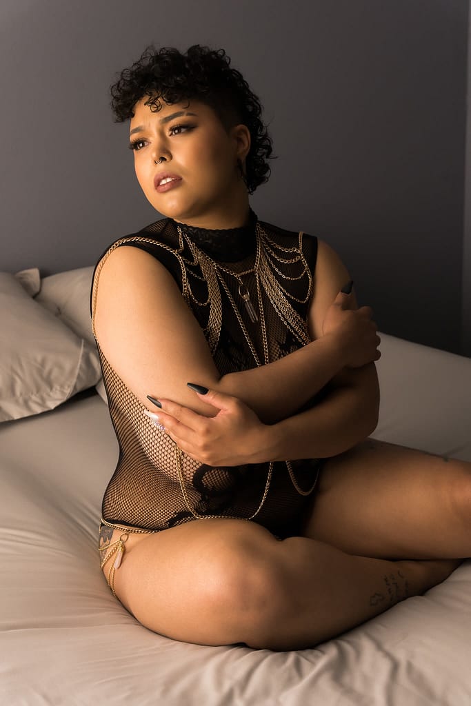 Women sitting on bed looking longingly and hugging her arms around her body, wearing a lacy bodysuit and gold body chain.  Photo by Embodied Art Boudoir. Denver boudoir photographer, denver boudoir photography, boudoir, colorado boudoir, denver boudoir, body positive, body positivity, body neutrality, body neutral, fat positive, haes, plus size, petite, body image, body love, self love, self compassion, mirror work, boudoir photos, sexy photos, mindset, beautiful bodies, healing body image