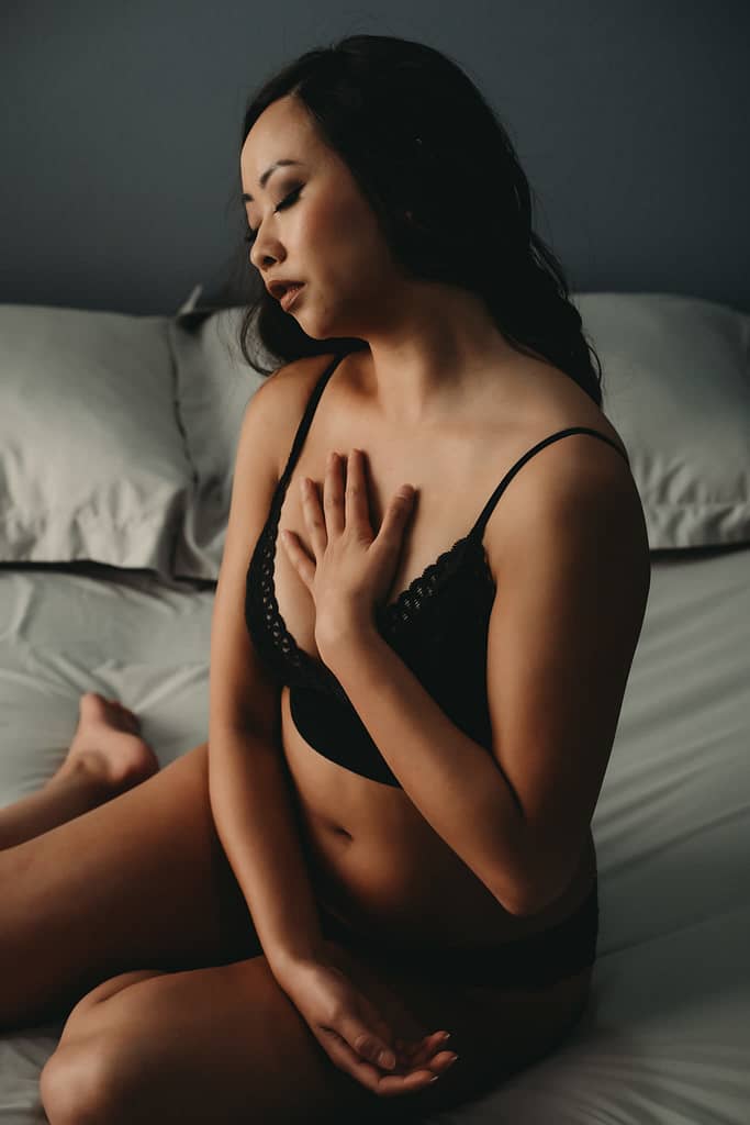 Woman sitting on bed, with her hand on heart and eyes closed wearing a matching black bralette and panties. Photo by Embodied Art Studio. Denver boudoir photographer, denver boudoir photography, boudoir, colorado boudoir, denver boudoir, body positive, body positivity, body neutrality, body neutral, fat positive, haes, plus size, petite, body image, body love, self love, self compassion, mirror work, boudoir photos, sexy photos, mindset, beautiful bodies, healing body image