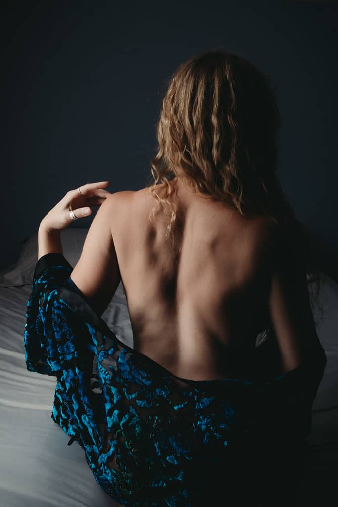 Woman sitting on bed, showing off back and curly blonde hair wearing a blue robe. Photo by Embodied Art Boudoir. Dark and moody, black and white photography, intense images, high intensity, memorable, sensual photography, sensual boudoir, colorado boudoir, denver boudoir, boulder boudoir, colorado springs boudoir, boudoir ideas, boudoir poses, boudoir inspiration, photography inspiration, romantic boudoir, bridal boudoir