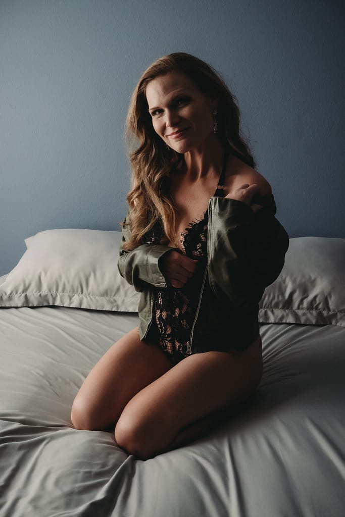 Blonde woman sitting on bed, smiling at camera wearing a lacy black bodysuit with a leather jacket. Photo by Embodied Art Boudoir. Intentions, intention setting, goal setting, boudoir photoshoot, boudior, buodoir, Denver boudoir, Colorado boudoir, boudoir photographer, self love, growth journey, affirmations, positive intentions, positive affirmations, self love journey, boudoir photography, Denver portrait photography, Denver portrait photographer, Boulder boudoir, fort collins boudoir, colorado springs boudoir, how to set an intention