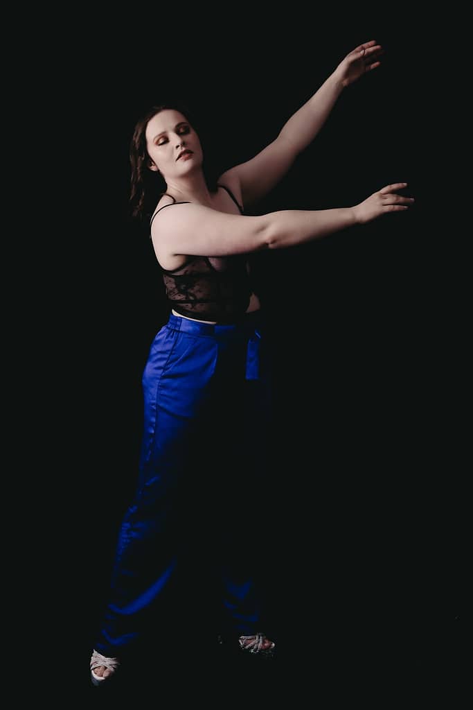 Woman standing in ballet like pose wearing shiny bright blue pants and a lacy top. Photo by Embodied Art Boudoir. Dark and moody, black and white photography, intense images, high intensity, memorable, sensual photography, sensual boudoir, colorado boudoir, denver boudoir, boulder boudoir, colorado springs boudoir, boudoir ideas, boudoir poses, boudoir inspiration, photography inspiration, romantic boudoir, bridal boudoir