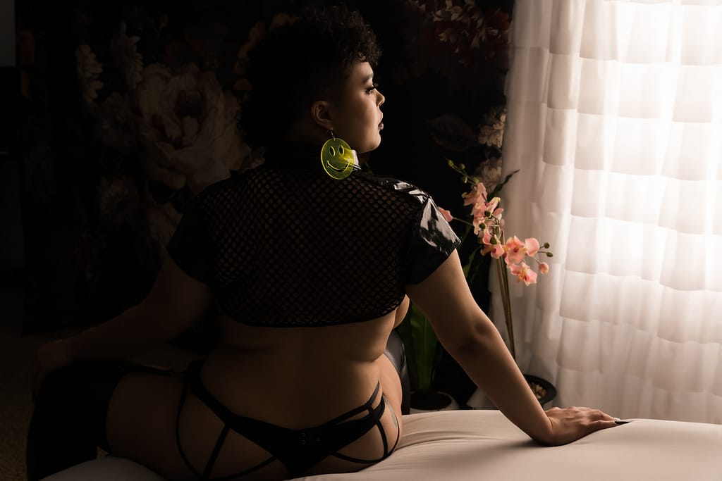A woman sits on the edge of a bed, looking toward the window. She's wearing strappy black lingerie and a has huge fluorescent  earring in the shape of a smiley face. Photo by Embodied Art Boudoir. Boudoir session, boudoir photoshoot, colorado boudoir, denver boudoir, self love, photoshoot, photoshoot packing list, packing list, boudoir packing list, what to pack, what to bring, boudoir outfits, boudoir inspo, outfit ideas, boudoir jewelry, shoes for boudoir