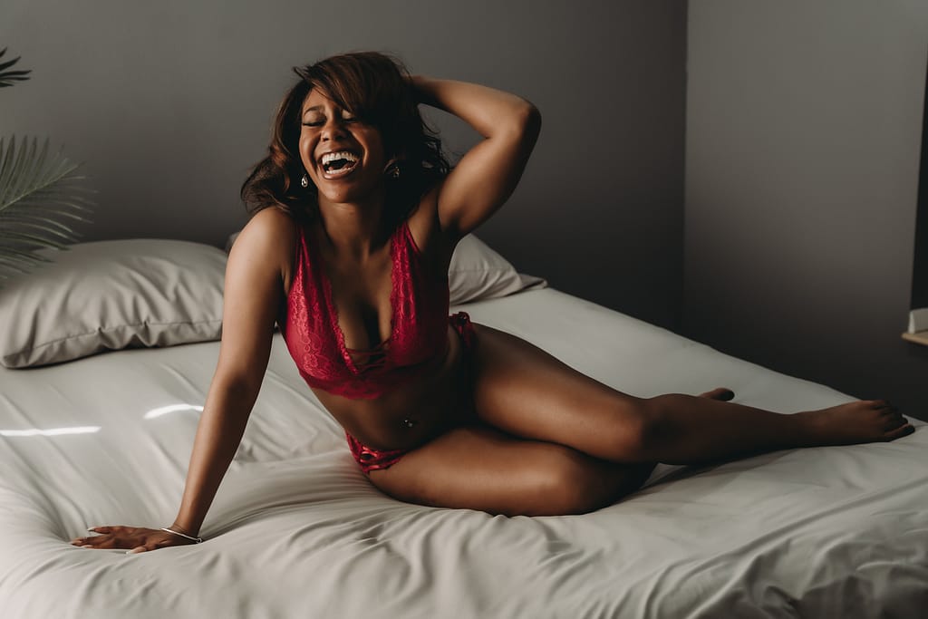 A woman sits on the bed, with a huge, happy laugh on her face! Her pink lacy bralette pops against her skin. Photo by Embodied Art Boudoir. Journaling, journal prompts, journal ideas, journal inspo, body image, body love, body appreciation, body positive, fat positive, boudoir, boudoir photos, boudoir session, boudoir photography, denver boudoir, colorado boudoir, boudoir inspo, boudoir ideas