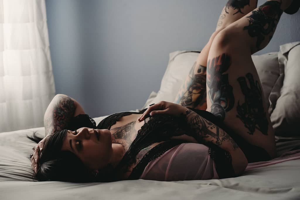 Plus sized tattooed woman lying on her back on bed with her hand on her chest wearing a lacy black bralette. Photo by Embodied Art Studio. Denver boudoir photographer, denver boudoir photography, boudoir, colorado boudoir, denver boudoir, body positive, body positivity, body neutrality, body neutral, fat positive, haes, plus size, petite, body image, body love, self love, self compassion, mirror work, boudoir photos, sexy photos, mindset, beautiful bodies, healing body image