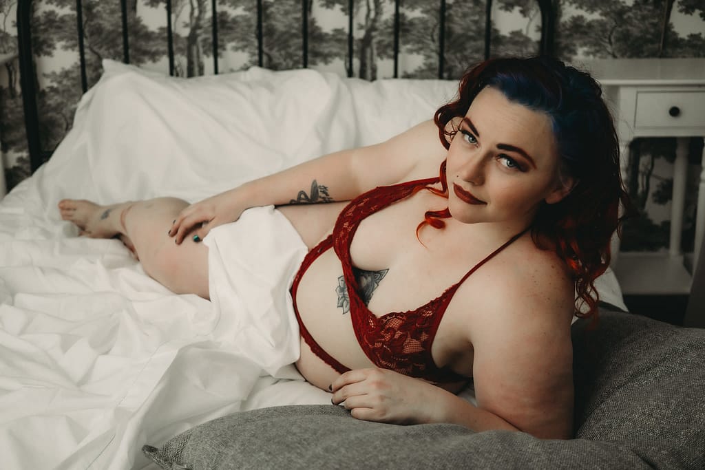 Woman lying on bed, looking at camera wearing a red bralette and matching panties. Photo by Embodied Art Boudoir, boudoir posing, boudoir inspiration, plus size photography, plus size boudoir, plus size glamour, plus size posing, plus size outfits, fat boudoir, fat empowerment, body positivity, fat positive, body positive, body confidence, body empowerment, haes, colorado boudoir, denver boudoir 
