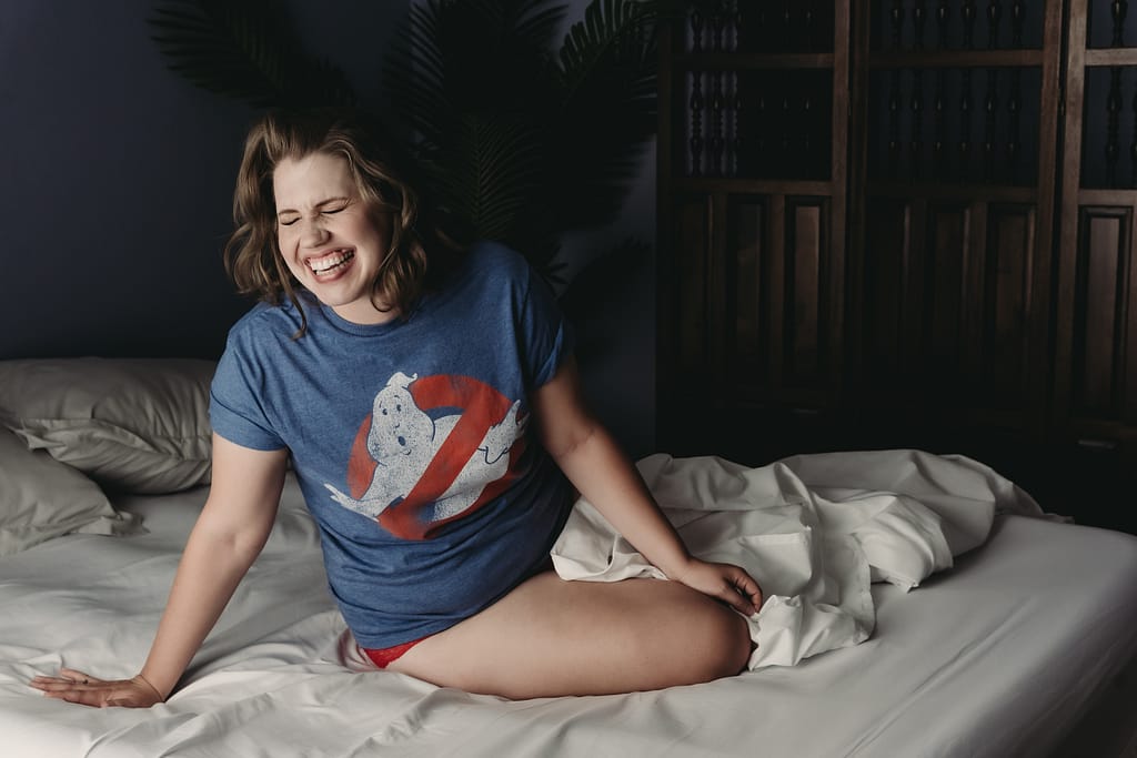 Blonde Woman laughing hard,  sitting on bed wearing a blue ghost busters tee shirt and  a white sheet. Photo by Embodied Art Boudoir. Empowering photography, boudoir photography, empowering boudoir, body positive, judgement free, body neutral, boudoir colorado, boudoir denver, empowerment, self love, self acceptance, safe space, boudoir photographer, classic boudoir, artistic boudoir, boudoir studio, studio photography, Empowering Boudoir Photography