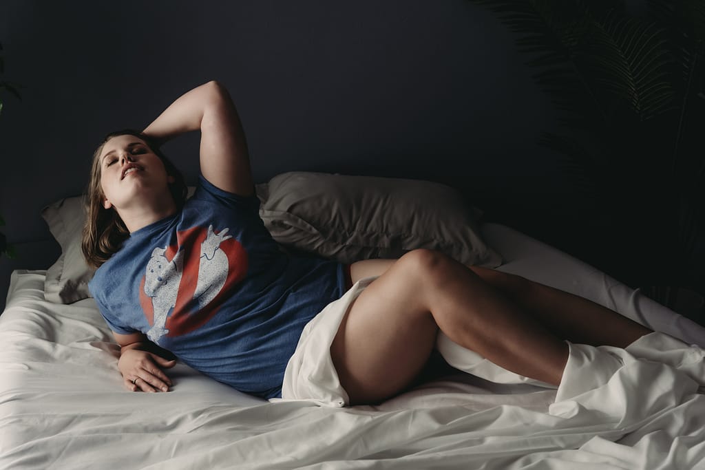 Women wears a ghostbusters t shirt during her boudoir session, posing on the bed with a pleased look on her face. Photo by Embodied Art Boudoir. Boudoir outfits, boudoir lingerie, boudoir fashion, boudoir babes, boudoir inspo, boudoir fashion, boudoir dress, boudoir style, personal style, favorite outfits, blogging, boudoir ideas, lingerie, colorado boudoir, boudoir, denver boudoir, denver boudior, boulder boudoir, colorado springs boudoir, boudoir ideas, photoshoot accessories, outfit inspiration, photoshoot outfit 
