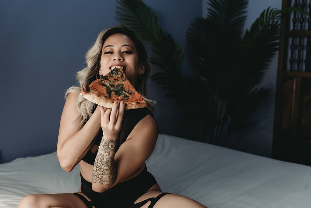 Woman sitting on bed, wearing black two piece enjoying a slice of pizza. Photo by Embodied Art Boudoir. Anti diet, health, body image, healthy body image, live well, body relationship, beautiful, self love, self-compassion, diet culture, sustainable practices, live well, female empowerment, media sensual boudoir, colorado boudoir, Denver boudoir, boulder boudoir, colorado springs boudoir, boudoir ideas, boudoir poses, boudoir inspiration, photography inspiration