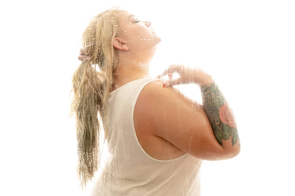 Woman standing in shower wearing a white tank top, holding her blonde hair. Photo by Embodied Art Boudoir. Body positive, body positive photography, fat positive, fat positive art, fat positive photography, colorado boudoir, denver boudoir, boulder boudoir, body positive colorado, haes, plus size photography, plus size art, women, beautiful, body positive art, big body art, you are beautiful, inclusive photography, inclusive art, celebrate all bodies, diverse photography