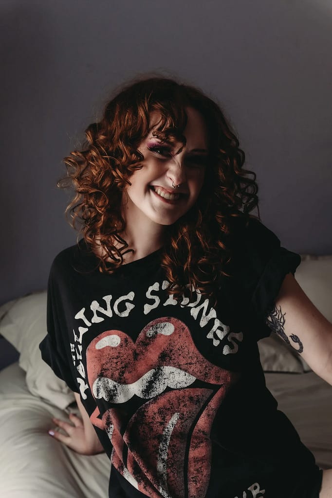 Red hair curly hair woman smiling sitting on the bed wearing a black tee. hoto by Embodied Art Boudoir, cozy, cozy boudoir, cozy vibes, cozy outfits, cozy and comfortable, cozy boudoir photography, cozy inspiration, keep it cozy, boudoir outfit, boudoir outfit inspiration, what to wear to boudoir, colorado boudoir, denver boudoir, boulder boudoir, colorado springs boudoir, boudoir ideas, boudoir poses, boudoir inspiration, photography inspiration