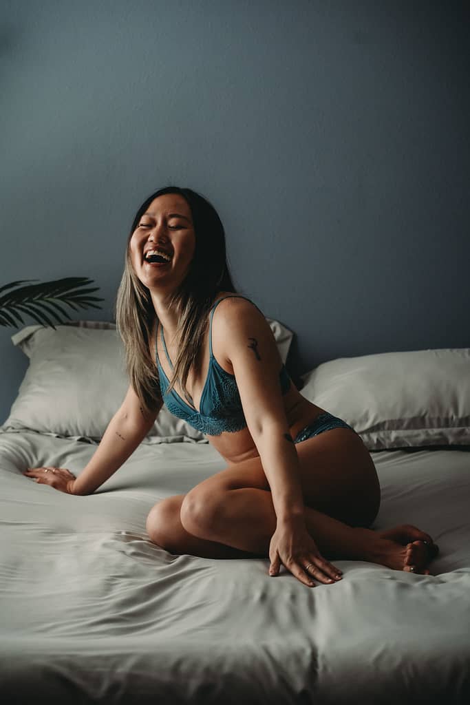 Woman sitting on bed laughing joyfully wearing a blue bralette with matching blue panties. Photo by Embodied Art Boudoir. Denver boudoir photographer, denver boudoir photography, boudoir, colorado boudoir, denver boudoir, body positive, body positivity, body neutrality, body neutral, fat positive, haes, plus size, petite, body image, body love, self love, self compassion, mirror work, boudoir photos, sexy photos, mindset, beautiful bodies, healing body image