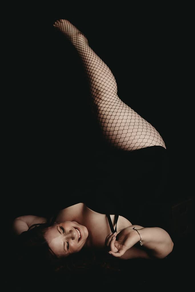 Woman lying down with legs in the air playfully smiling. Wearing a black body suit and fishnet stockings. Photo by Embodied Art Boudoir. Boudoir photography, self-care, journal, journal inspiration, journaling, journal addict, journal lover, journal inspo, self-care tips, self-care first, self-care routine, self-care tips,  self-care daily, healthy choices, healthy habits, habits, success habits, habits for success, me time, quotes to live by, colorado, colorado photographer, Denver boudoir, Denver boudior, Colorado boudior, Embodied art boudoir, boudior,  boudoir, boudoir photo, photographer
