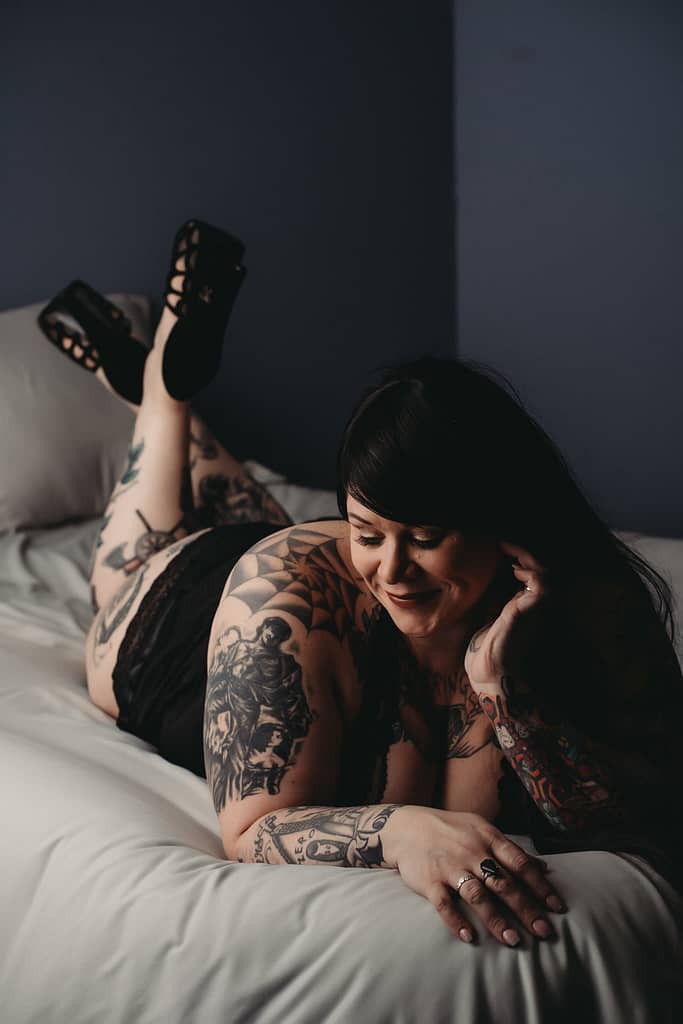 Woman lying on bed with many tattoos, wearing a black negligee and heels.Photo by Embodied Art Boudoir, boudoir posing, boudoir inspiration, plus size photography, plus size boudoir, plus size glamour, plus size posing, plus size outfits, fat boudoir, fat empowerment, body positivity, fat positive, body positive, body confidence, body empowerment, haes, colorado boudoir, denver boudoir 