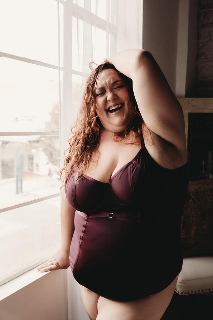 Woman standing joyfully next to a window wearing a deep purple body suit. Photo by Embodied Art Boudoir. Curvy women, curvy girl style, plus size boudoir, plus size photography, plus sized women, fight the inner critic, self confidence, self love, love your body, stylish plus sized women, plus size lingerie, beautiful women, empowered women, photogenic, stunning boudoir images, boudoir imagery, boudoir picture, female photographer, denver boudoir, golden boudoir, golden colorado boudoir, boudoir inspiration 