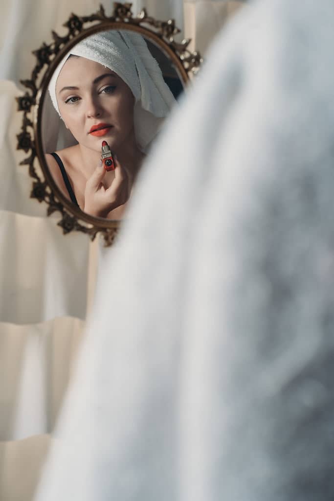 Woman looking softly into an antique mirror with a towel on her head, applying red lipstick. Photo by Embodied Art Boudoir. Boudoir photography, boudoir photos, denver boudoir, colorado boudoir, denver boudoir photographer, pinup, pin up, pin-up, boudoir studio, boudoir session, sensual photography, fine art nude photography, fine art nudes, boudoir, boudoir history, photography history, 70s photography, 70s photoshoot 