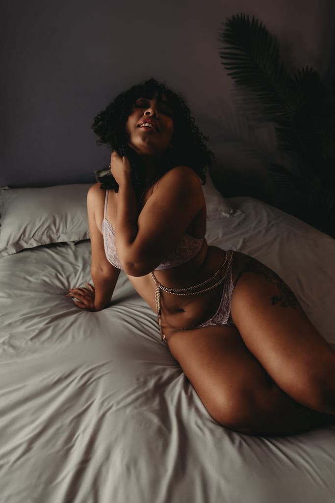 Black woman lying on bed wearing a purple bralette and matching panties with a gold chain. Photo by Embodied Art Boudoir. Boudoir photography, boudoir photoshoot, photoshoot, photo book, photo album, velvet cover, velvet album, boudoir album, black album, professional album, professional printer, wall art, prints, folio box, folio, collection, nude art, photography, golden boudoir, colorado boudoir photographer, denver boudoir photographer