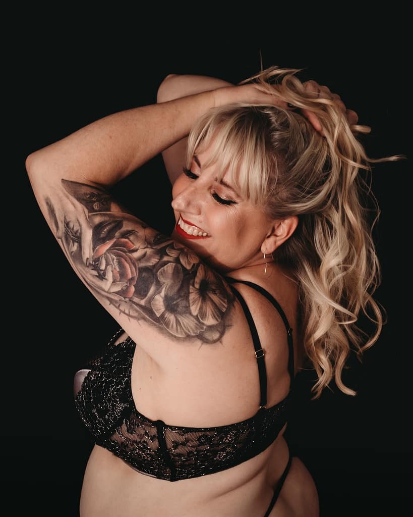 Blonde woman holding her hair up, smiling to the side, showcasing her arm tattoo wearing a black bra and matching thong. Photo by Embodied Art Boudoir. Feeling safe, feeling secure, security, safety, comfortable, professional photographer, female photographer, confidence, self-confidence, personal growth, colorado boudoir, denver boudoir, boulder boudoir, colorado springs boudoir, boudoir ideas, boudoir poses, boudoir inspiration, photography inspiration